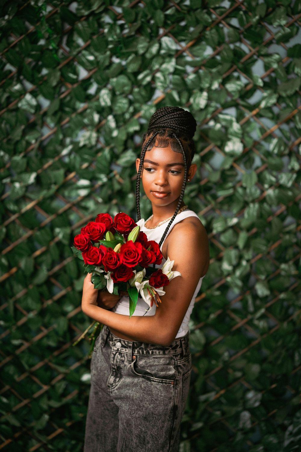 Fashion portrait with red roses against lush green foliage backdrop wearing white tank top and acid wash jeans.
