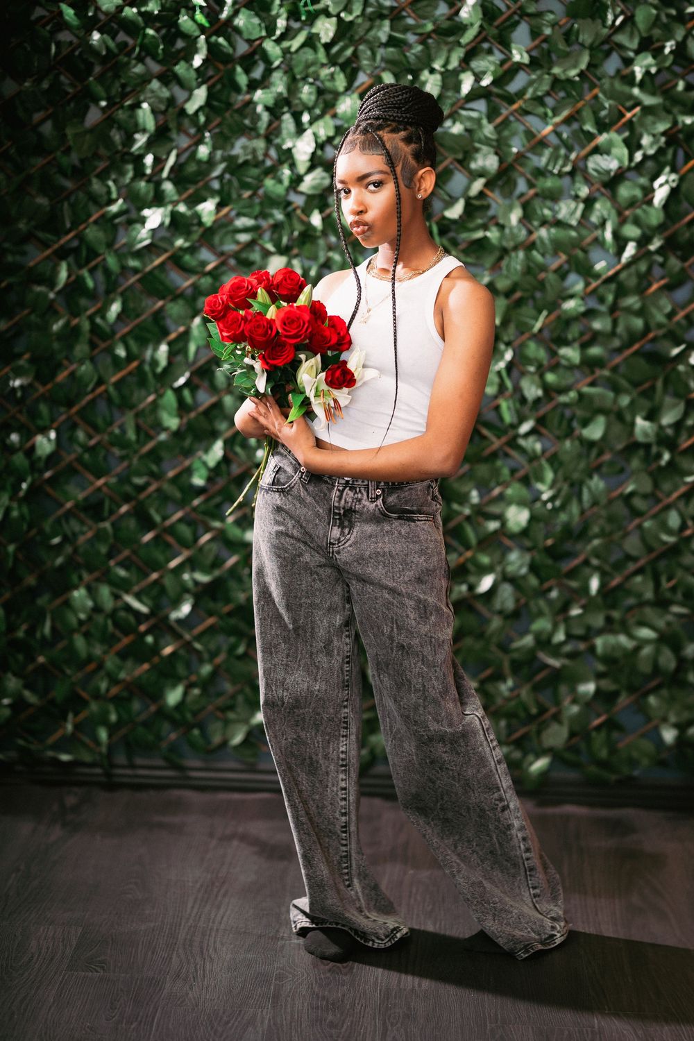 Fashion portrait of model in white tank top and jeans holding bouquet against green wall.