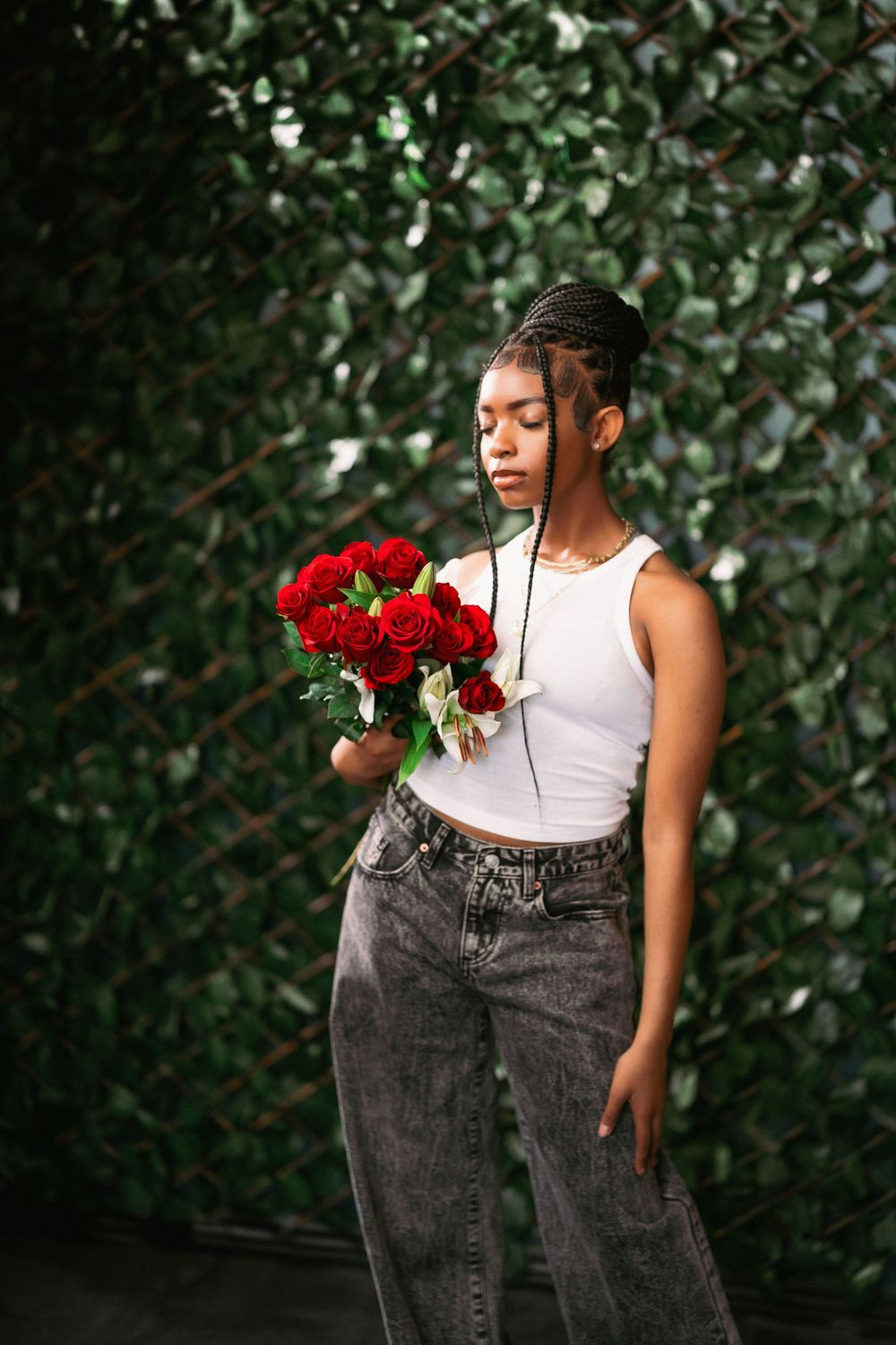Artistic portrait of model posing with red roses against verdant wall backdrop.