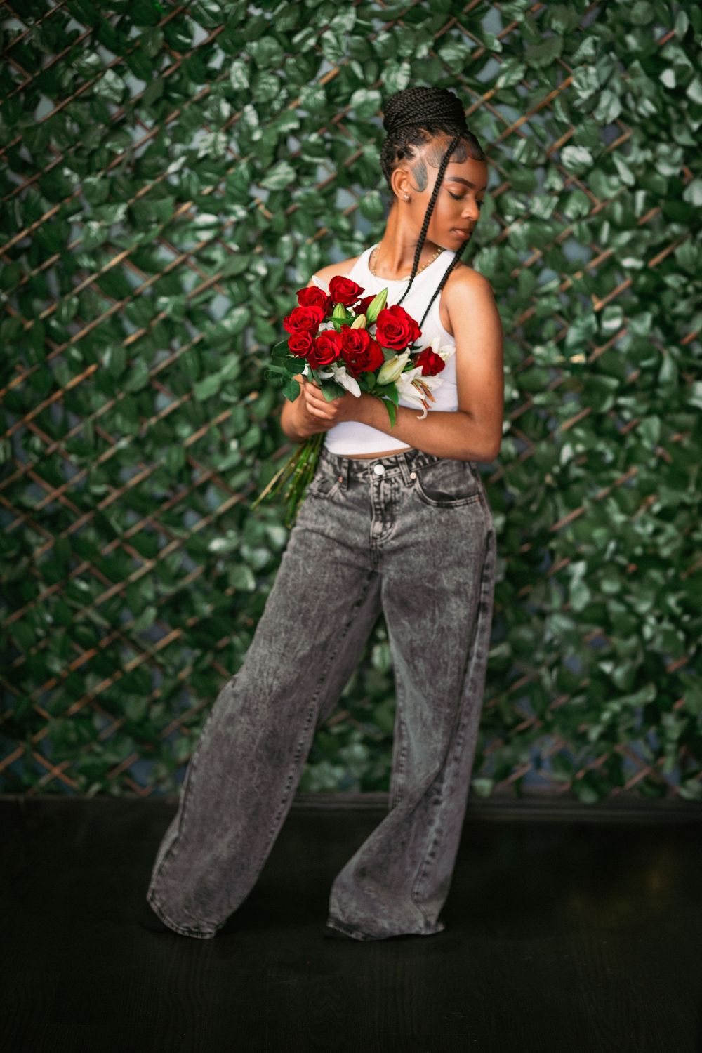 Creative portrait pose with red roses against leafy backdrop wearing casual denim and white tank top.