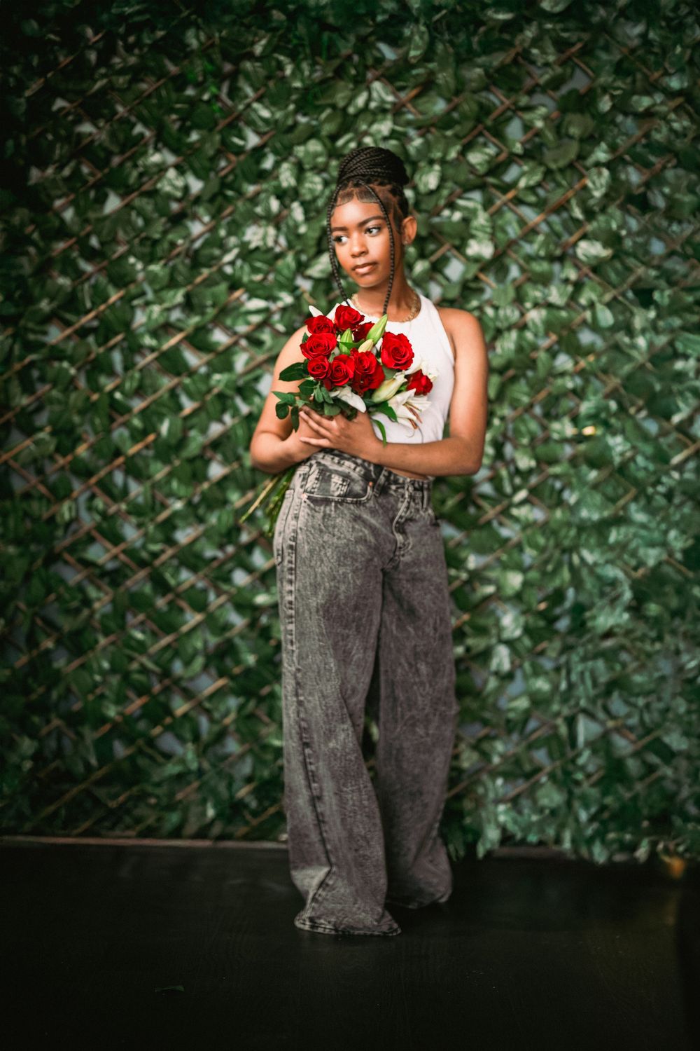 Full-length fashion portrait with red roses against green wall wearing white top and wide-leg jeans.