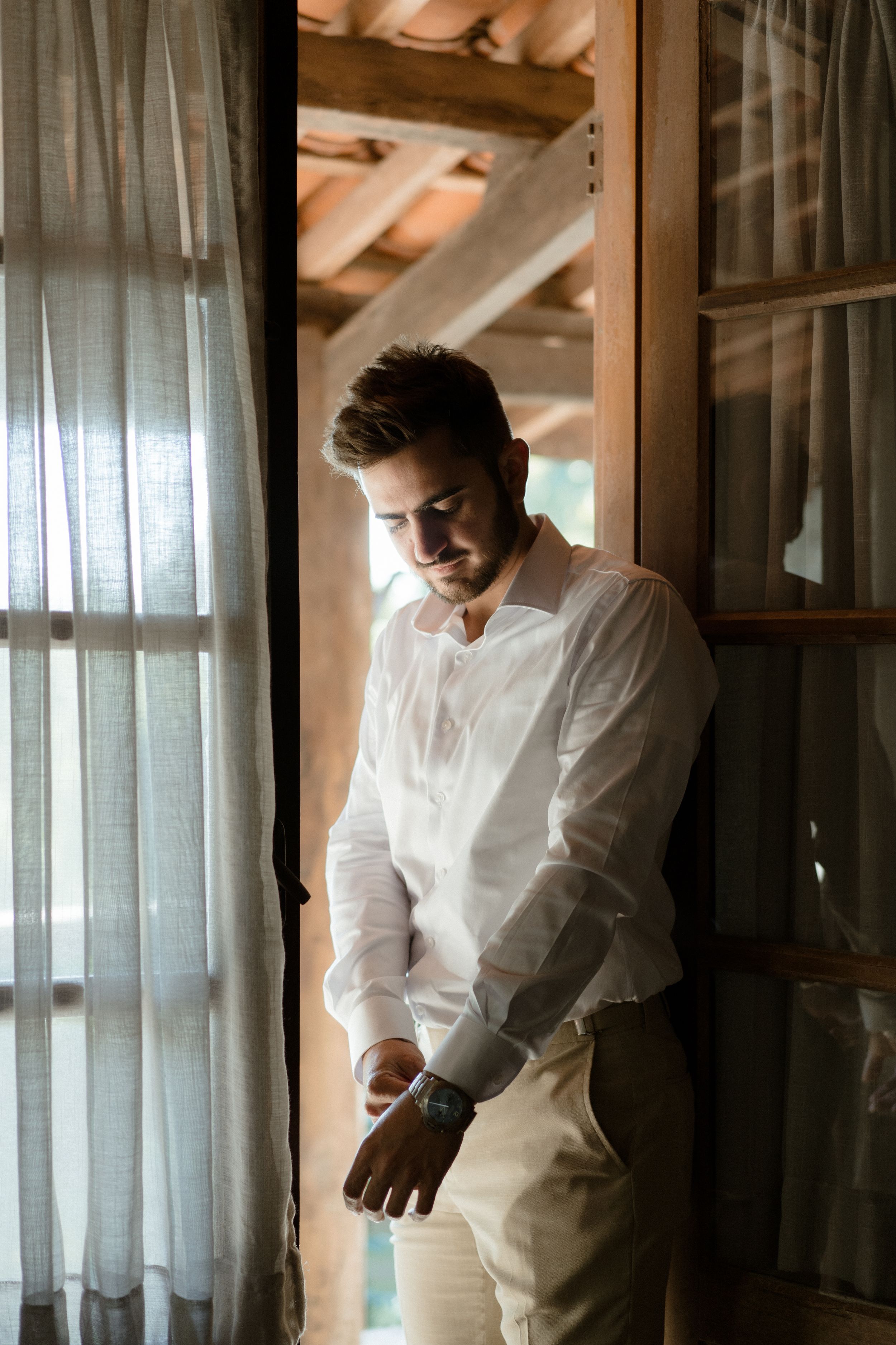 A person in a white shirt and khaki pants stands by a curtained window in natural light.
