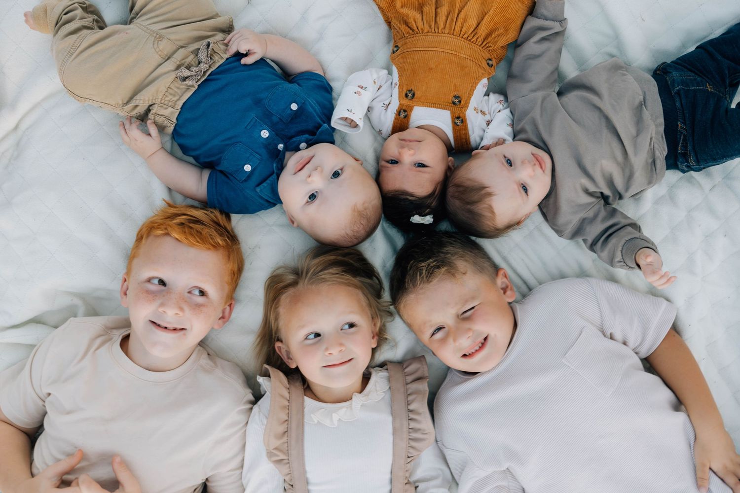 Five young children lying in a circle on a white blanket, smiling up at the camera.