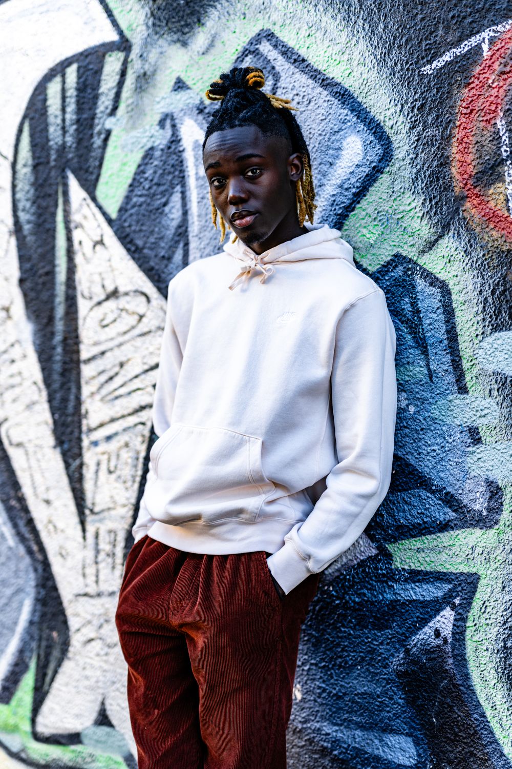 Person in white hoodie and corduroy pants poses against colorful graffiti wall.