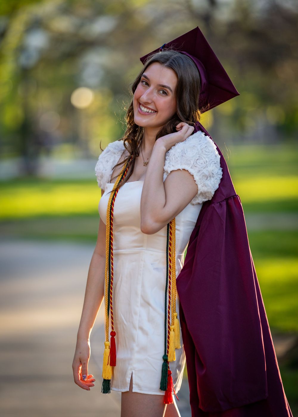 Graduate in maroon cap and gown with honor cords poses outdoors.