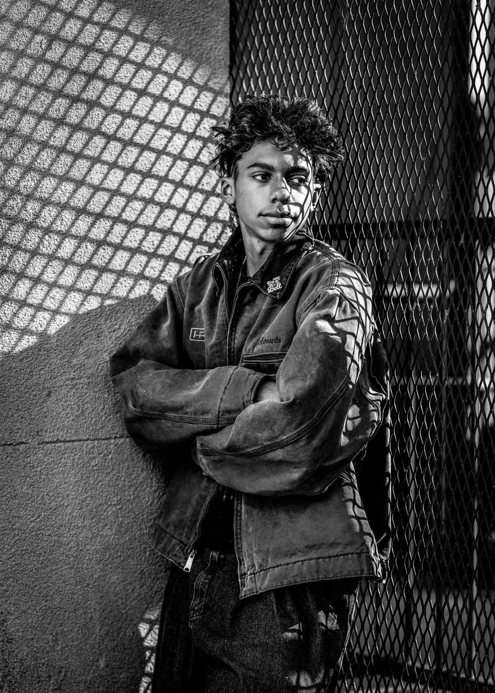 A dramatic black and white portrait of someone in a jacket leaning against a wall with mesh fencing in the background.
