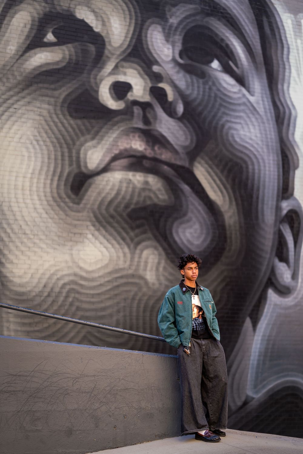 A person in a teal jacket poses against a dramatic black and white wall mural with swirling abstract patterns.