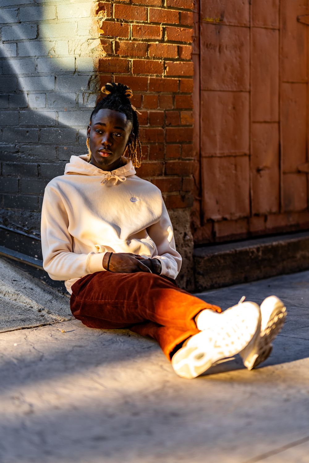 Person in white hoodie and orange pants sits against brick wall in sunlight.