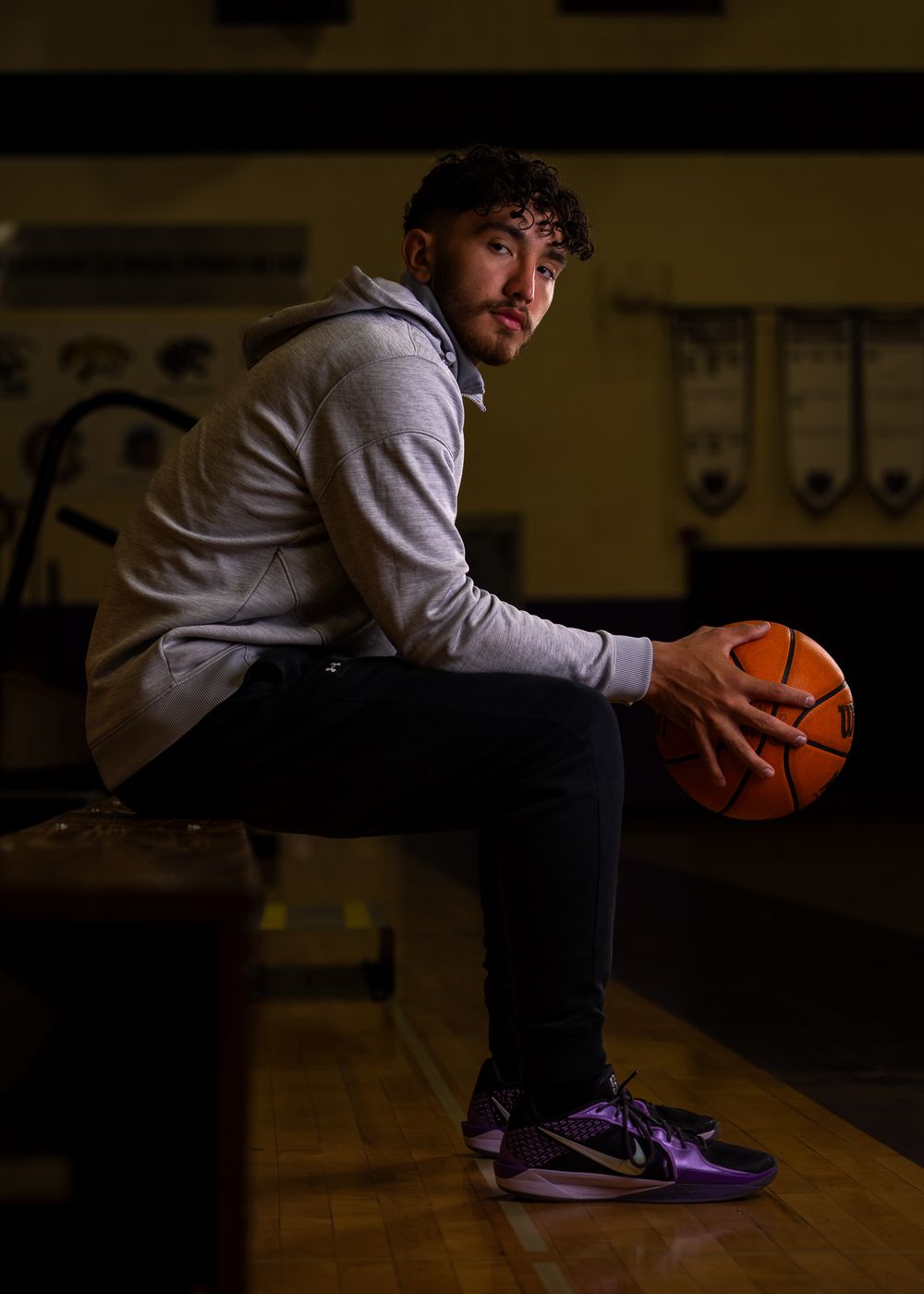 Basketball player in gray hoodie sits holding ball in dimly lit gymnasium.