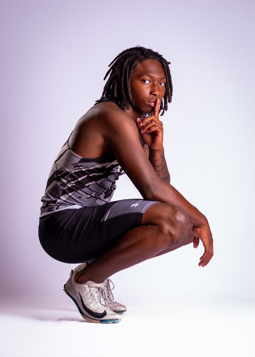 Athletic person in gray tank top and black shorts crouches in dynamic pose against light purple backdrop.