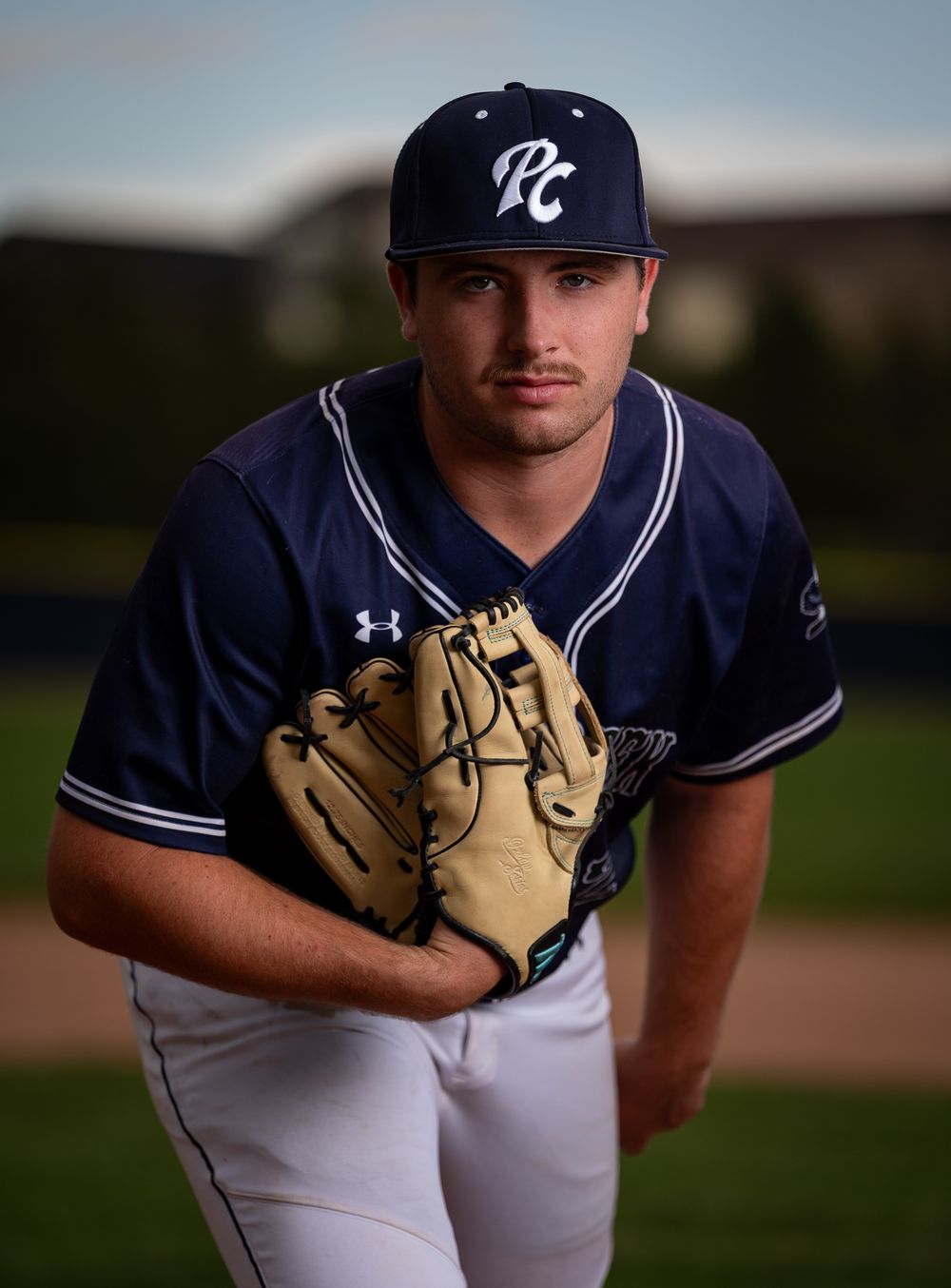 Baseball player in navy blue uniform holds up leather glove while posing for portrait on baseball field.