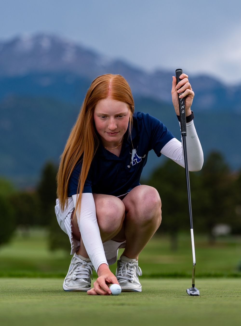 Golfer crouches to line up a putt on a scenic mountain golf course green.