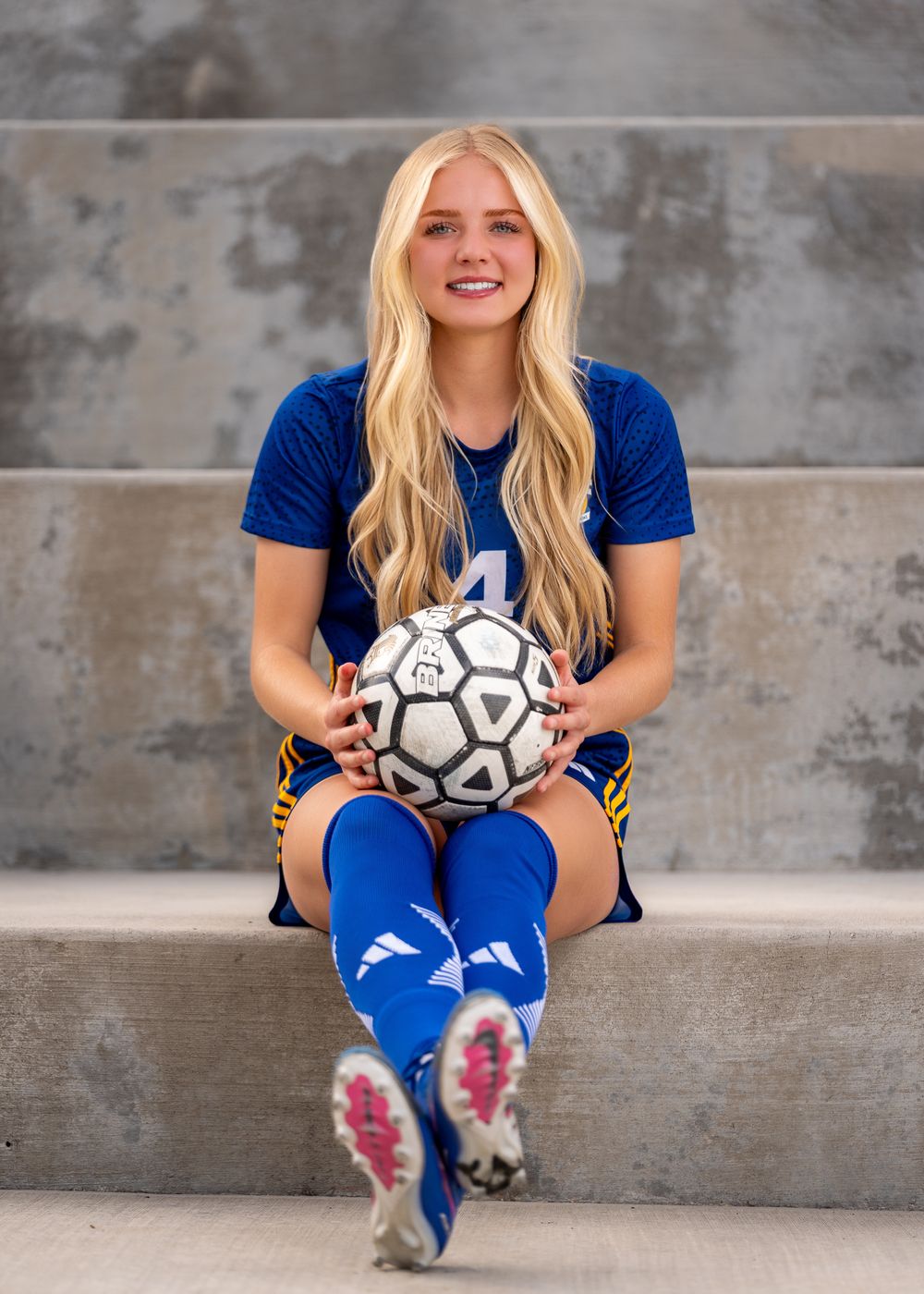 Young female soccer player in blue uniform sitting on bleachers holding a soccer ball, smiling for a team photo.