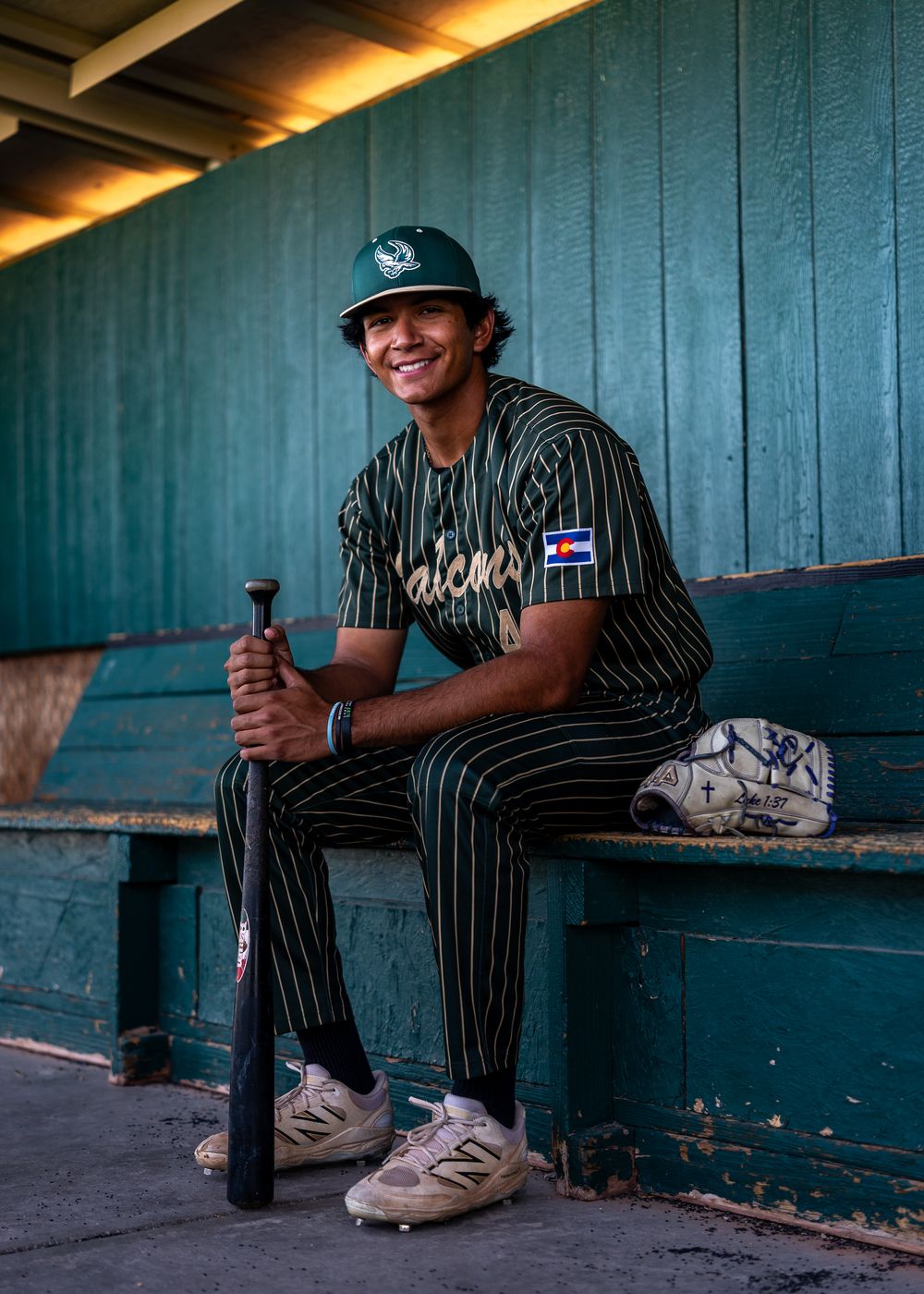 Young baseball player in green pinstripe uniform smiling in dugout, holding bat, with glove beside him.