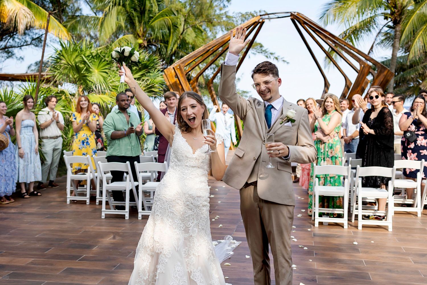 A couple in formal wedding attire walks across a wooden bridge surrounded by lush green trees in a park setting.