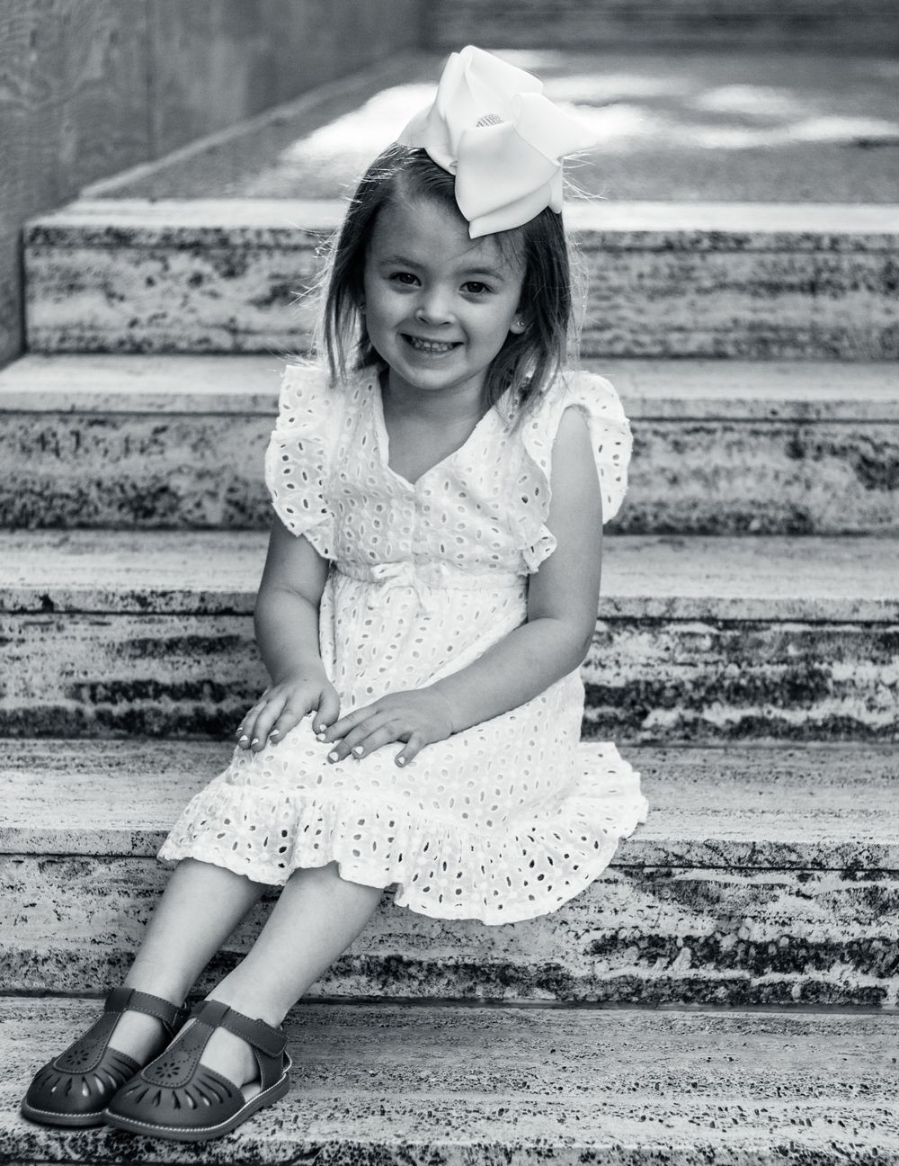 Black and white portrait of a happy young child in an eyelet lace dress sitting on stone steps with a bow in their hair.