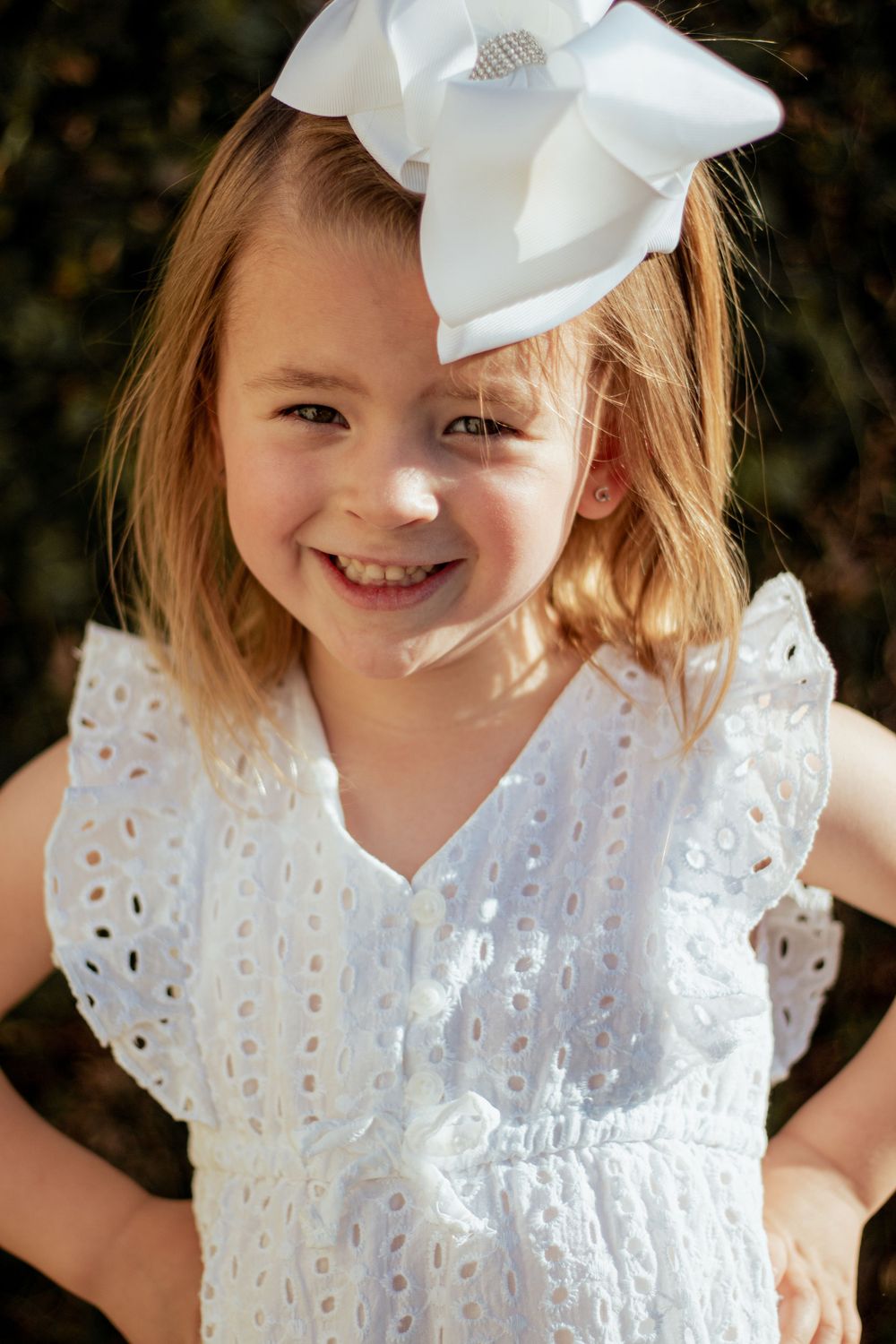 Young child wearing white eyelet dress and large white bow smiles brightly in outdoor portrait.