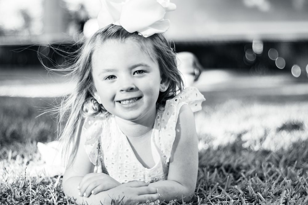 A joyful child in a white lace dress lies in the grass, captured in black and white photography.