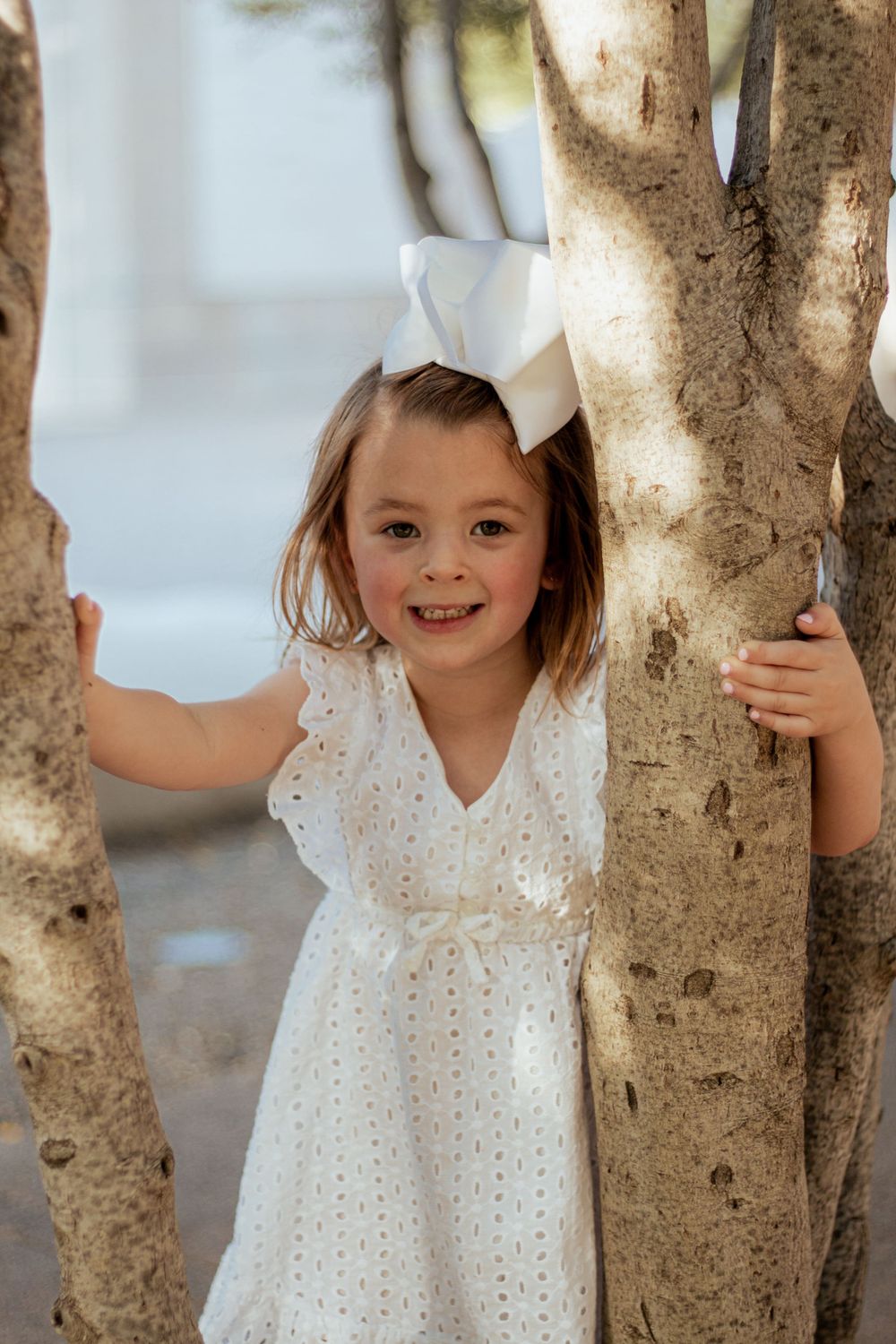 A young child in a white eyelet dress and bow stands between tree trunks on a sunny day.