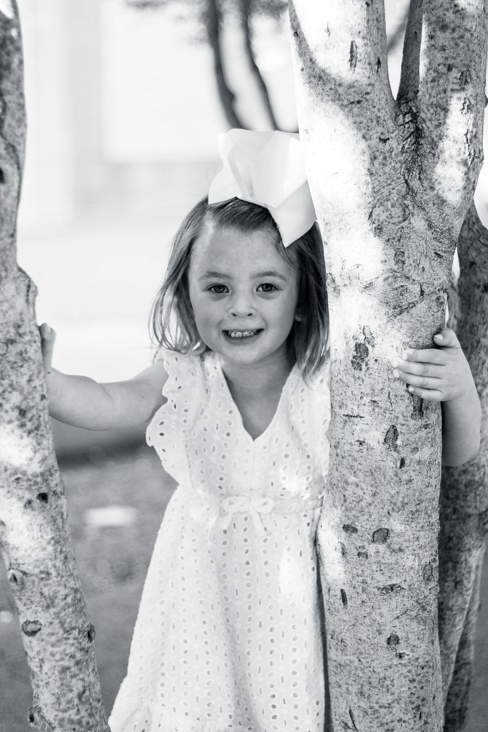 Black and white portrait of a child in a white dress standing between two tree trunks outdoors.