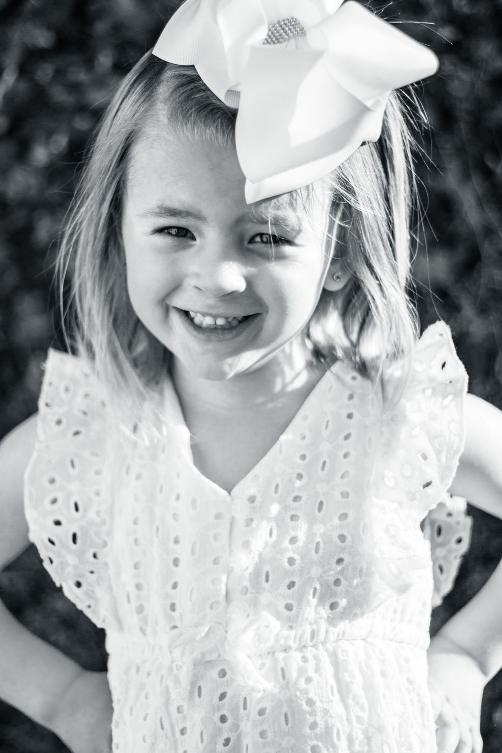 Black and white portrait of a child in white eyelet dress smiling with a large white bow in their hair.