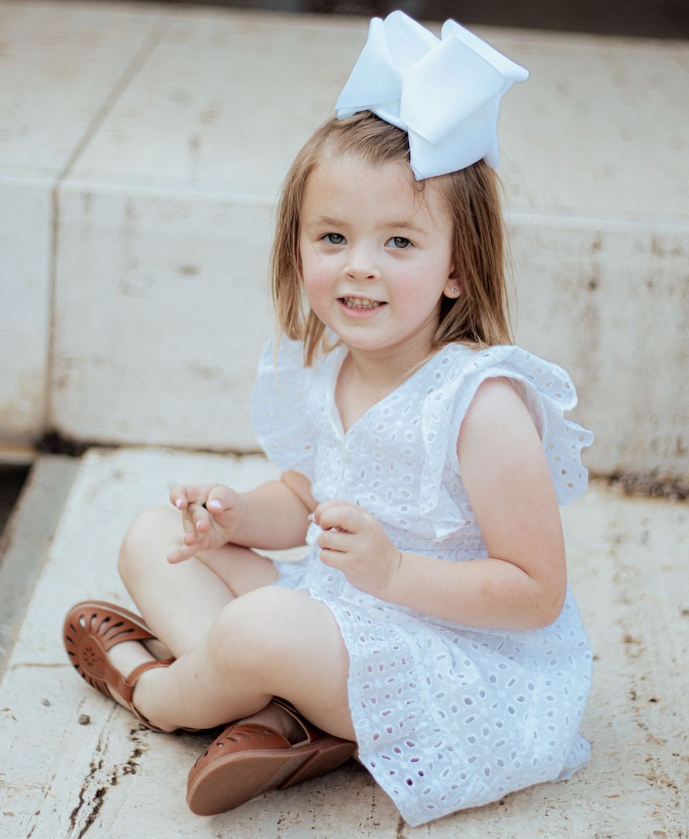 Small child sitting in white eyelet lace dress and brown sandals with large white bow hair accessory.