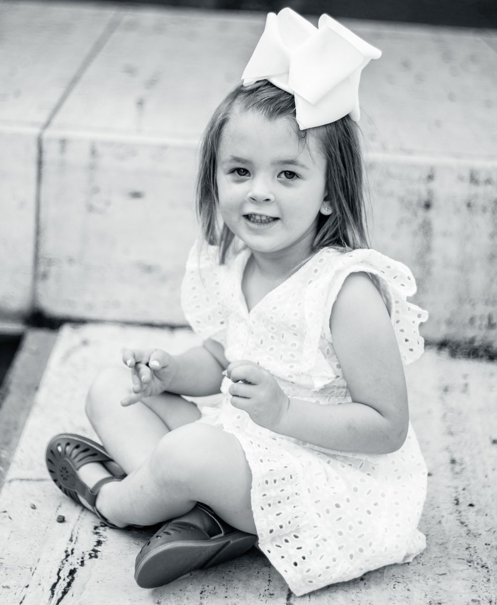 A young child in a white eyelet dress and hair bow sits on concrete steps in a classic black and white portrait.