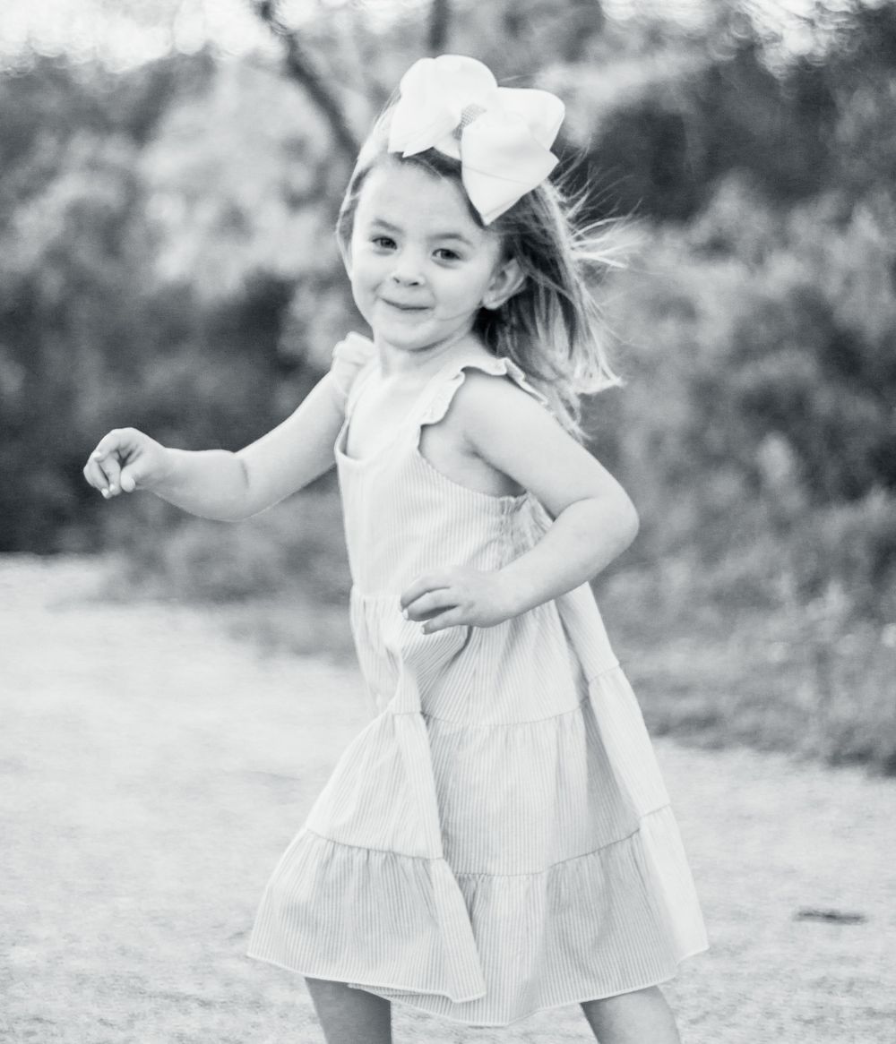 Black and white candid shot of a child in a white dress and bow dancing outdoors with carefree energy.