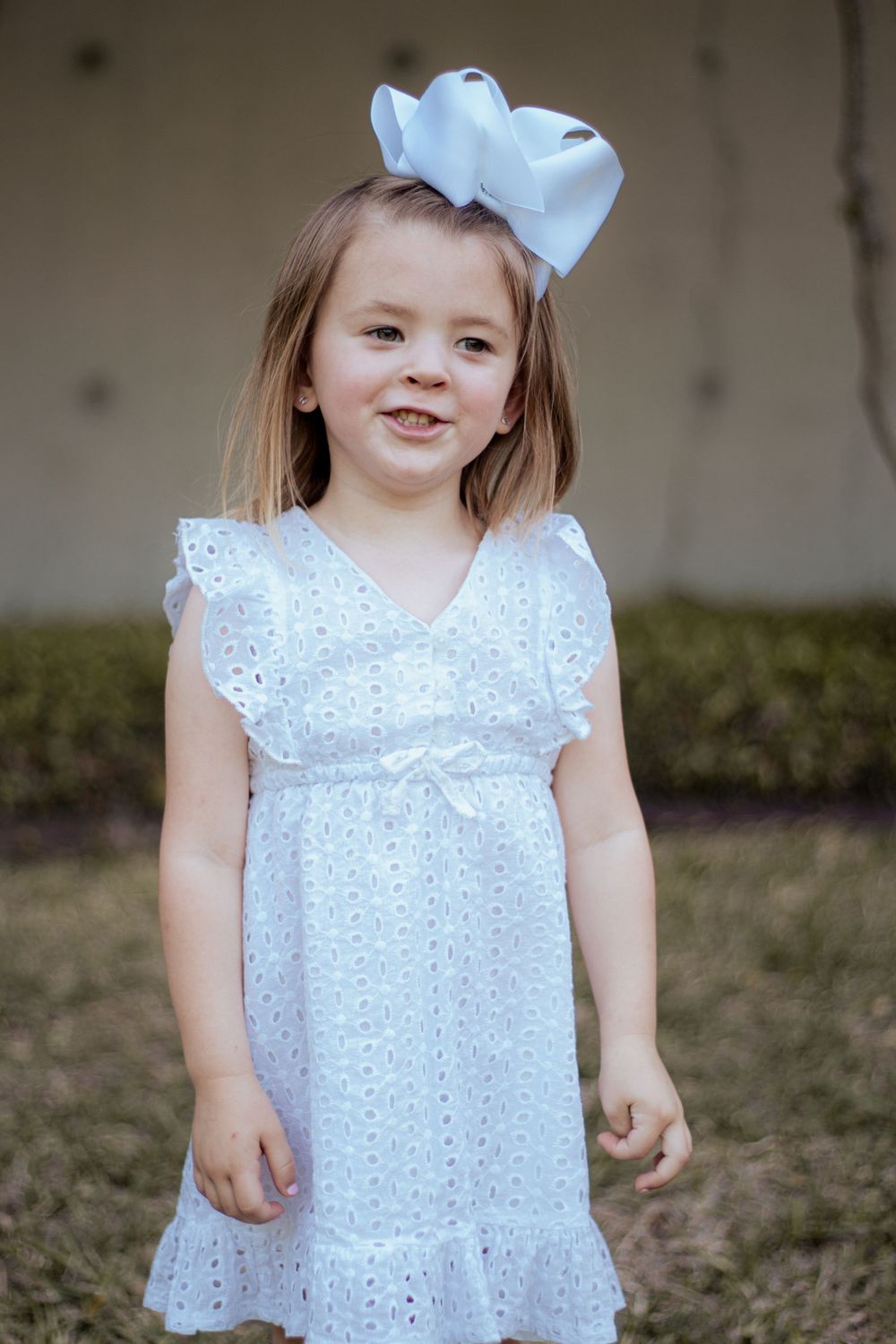 Young child wearing a white eyelet lace dress with flutter sleeves and a matching bow hair accessory in an outdoor setting.