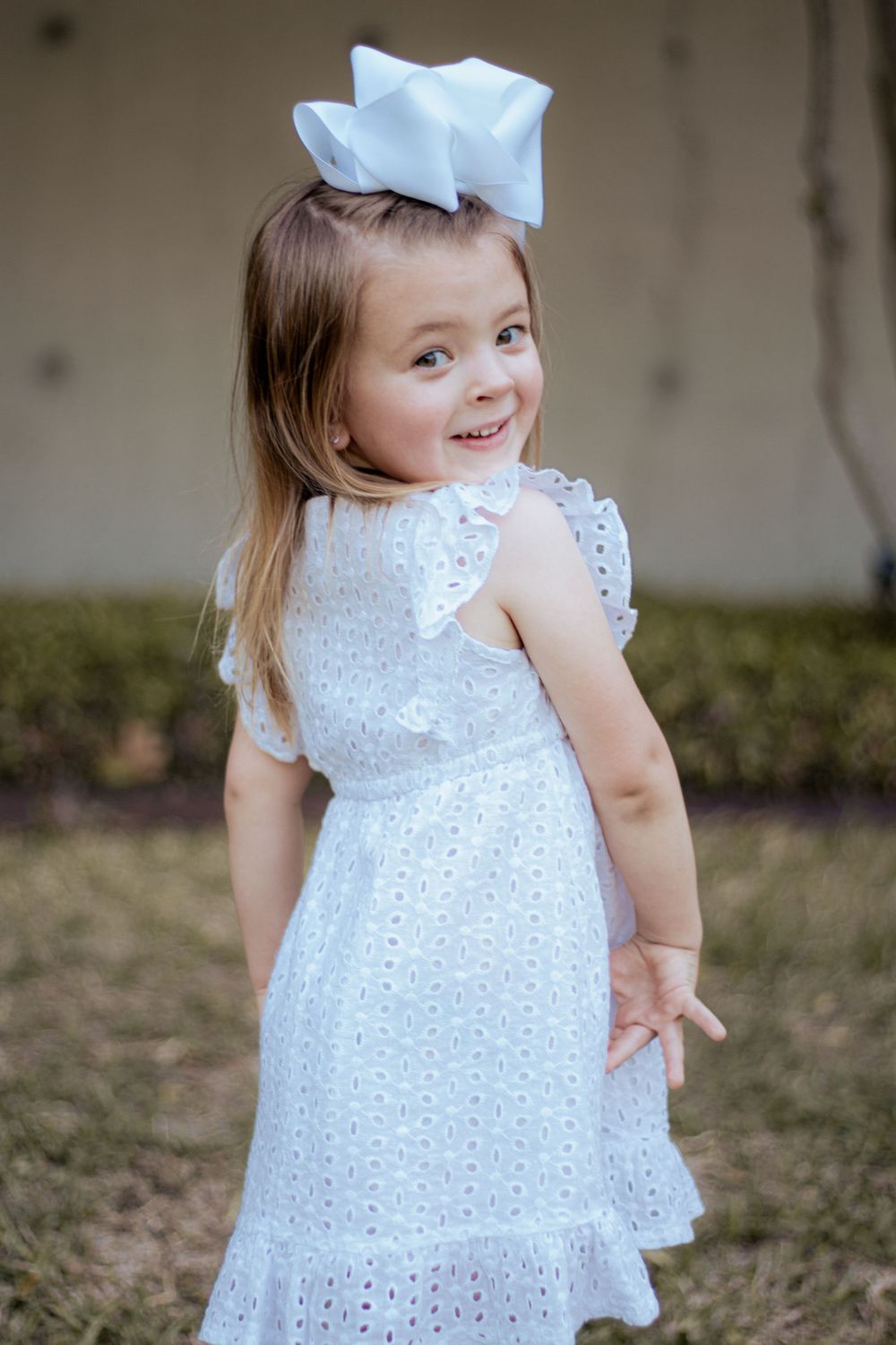 A small child wearing a white eyelet lace dress and large blue bow twirls outdoors with a playful expression.