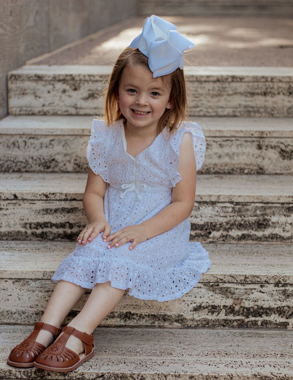 Child in white lace dress and bow sits smiling on concrete steps wearing brown leather sandals.