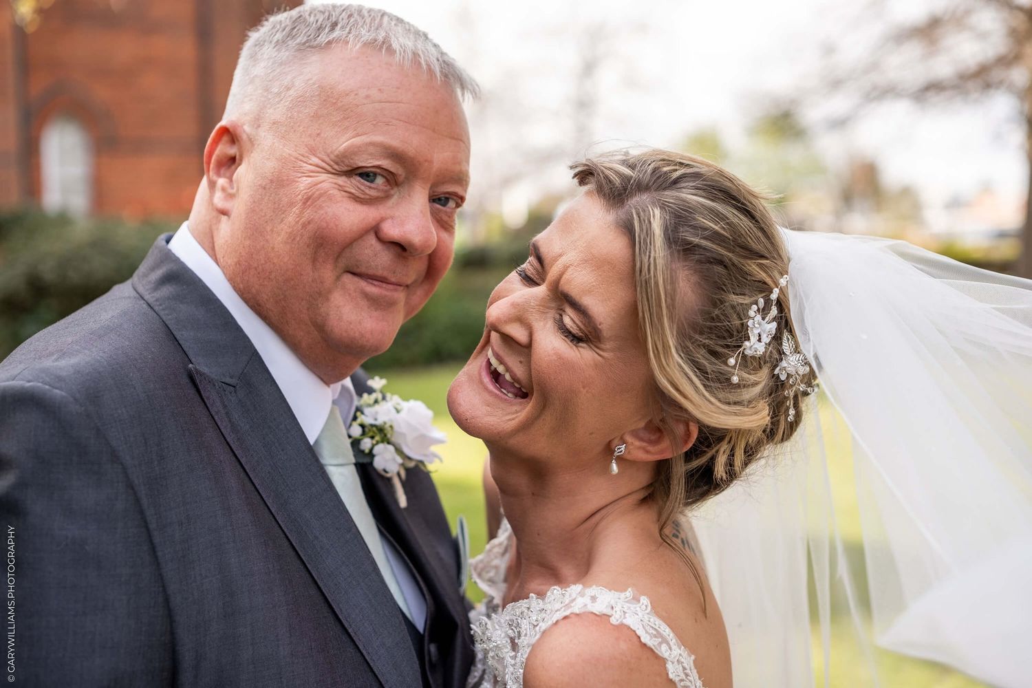 A happy couple shares a tender moment outdoors on their wedding day, the bride wearing a lace veil and floral headpiece.