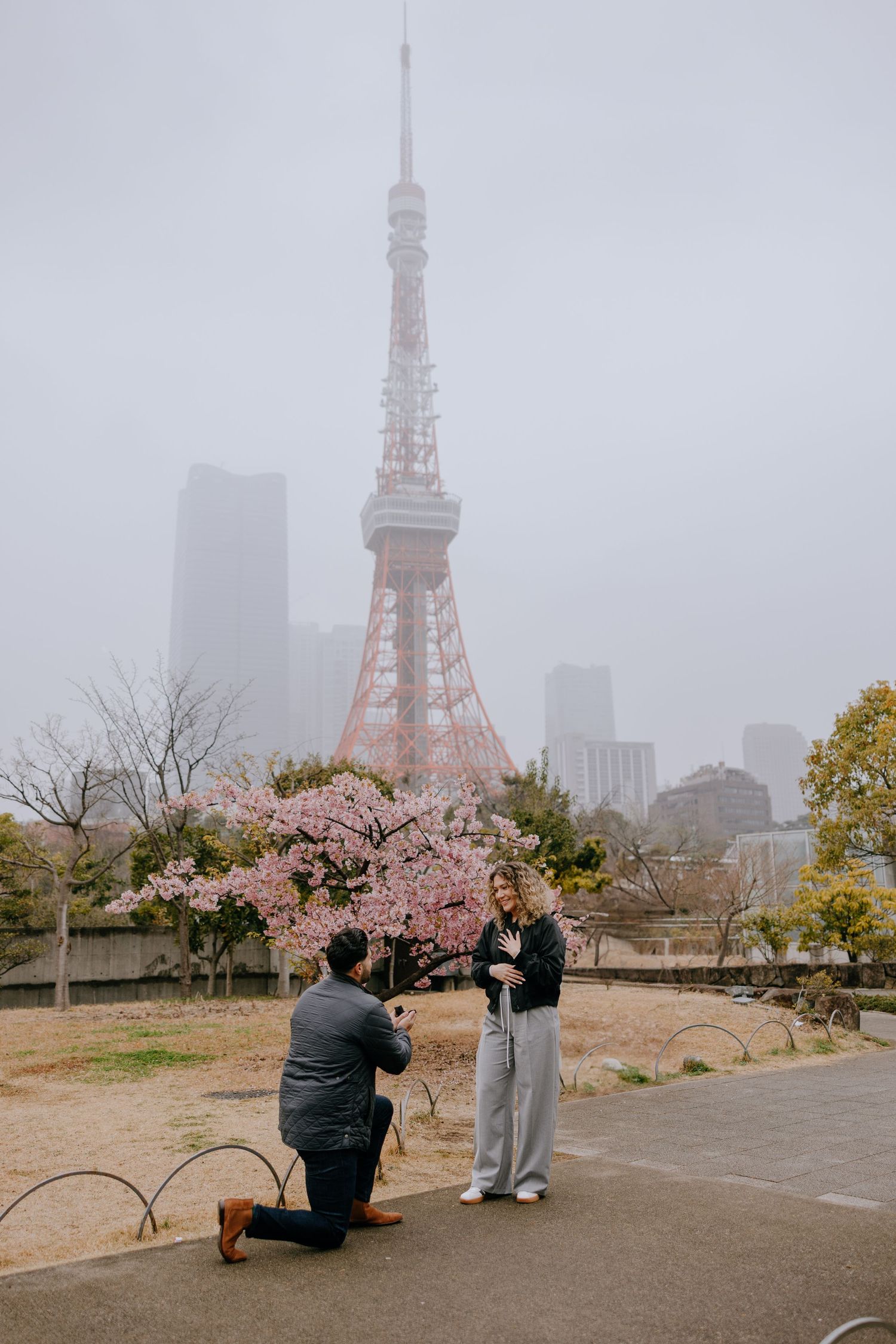 A romantic proposal moment at Tokyo Tower during cherry blossom season with foggy cityscape in the background.