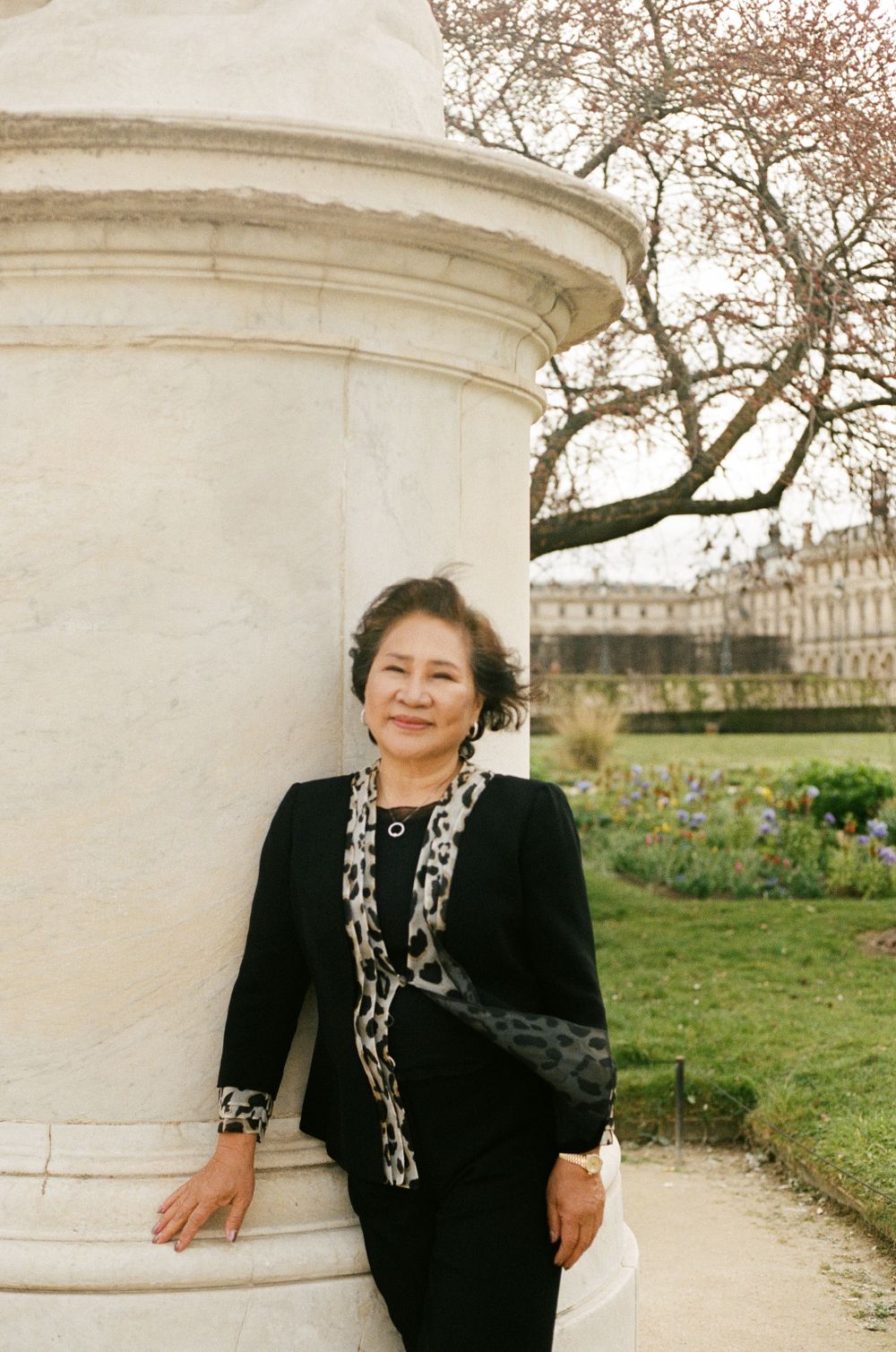 A woman in a black blazer with a patterned scarf stands beside a white stone column with a palace in the background.