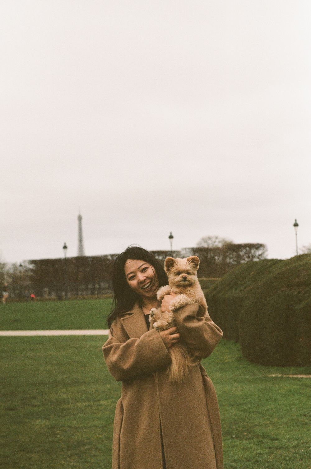 A person in a tan coat holds a small Pomeranian dog with the Eiffel Tower visible in the distant background in Paris.