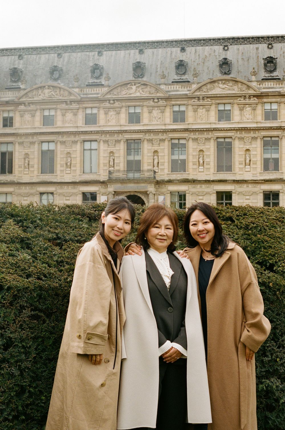Three people in warm coats standing together in front of an ornate European palace building with classical architecture.
