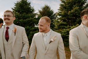 Groomsmen in light colored suits pose together against blue sky and evergreen trees.