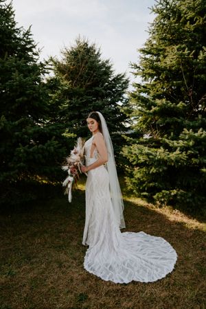 Bride in lace gown with flowing train poses among evergreen trees.