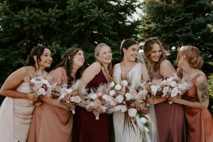 Bridesmaids in dusty rose dresses pose together holding white floral bouquets at an outdoor wedding.