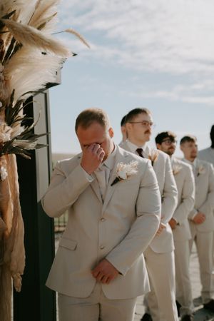Groomsmen in light beige suits stand in line during outdoor wedding ceremony.