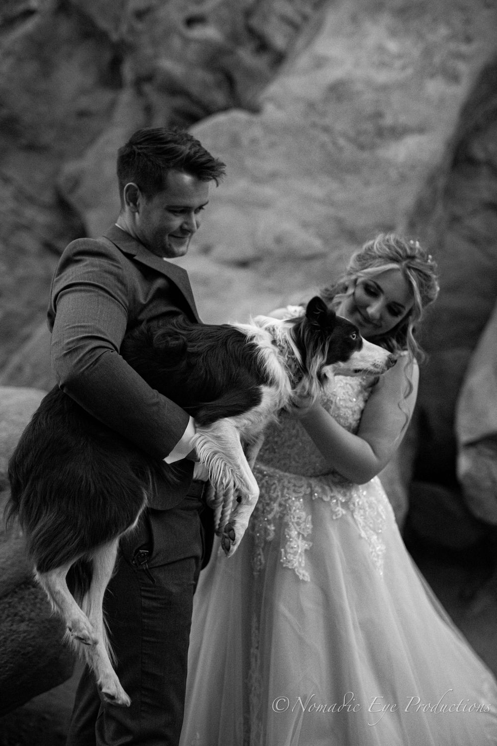 A couple pets their black and white dog during their wedding photoshoot among red rocks.