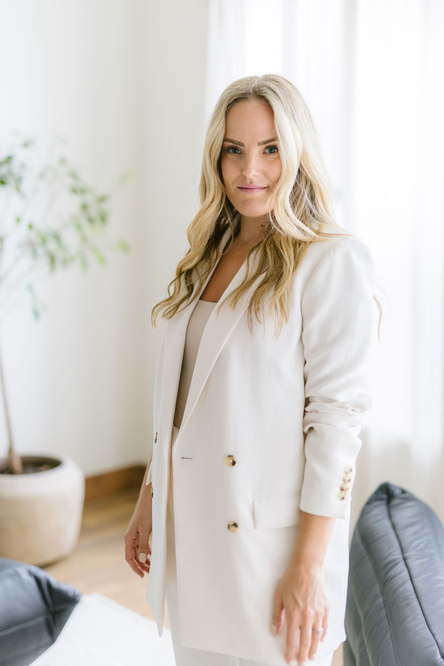 Professional portrait of someone wearing a crisp white blazer and blonde wavy hair in a bright modern interior setting.