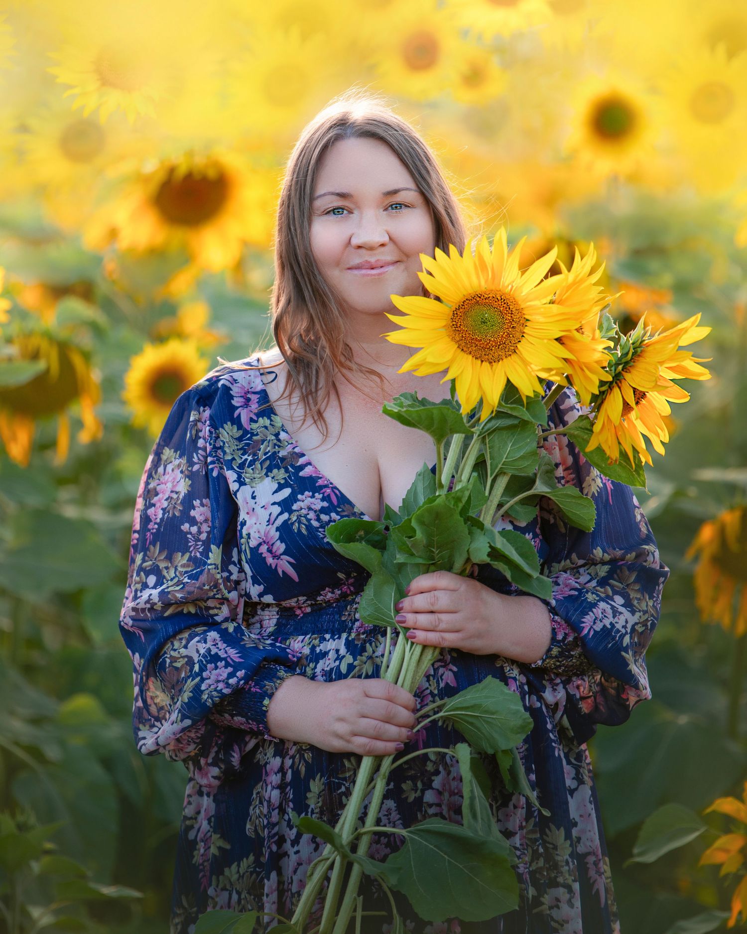 Someone in a blue floral dress holds freshly picked sunflowers while standing in a vibrant sunflower field at sunset.