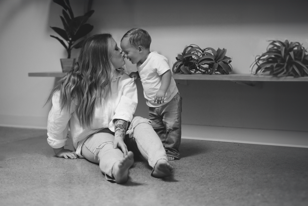 Black and white image of adult and small child playing together on a carpeted floor near indoor plants.