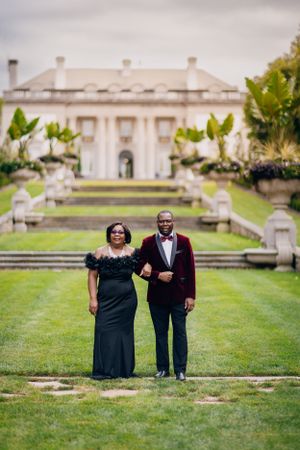 A couple poses in formal wear in front of a grand mansion with manicured gardens.