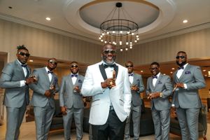 Wedding party in coordinated gray suits and white jacket stands together in an elegant room with circular chandelier.