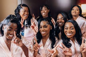 Bridal Party wearing pink satin robes smile together while making peace signs during a celebration photoshoot.