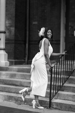 Black and white photo of a smiling woman in white dress and heels posing on outdoor steps with railing.