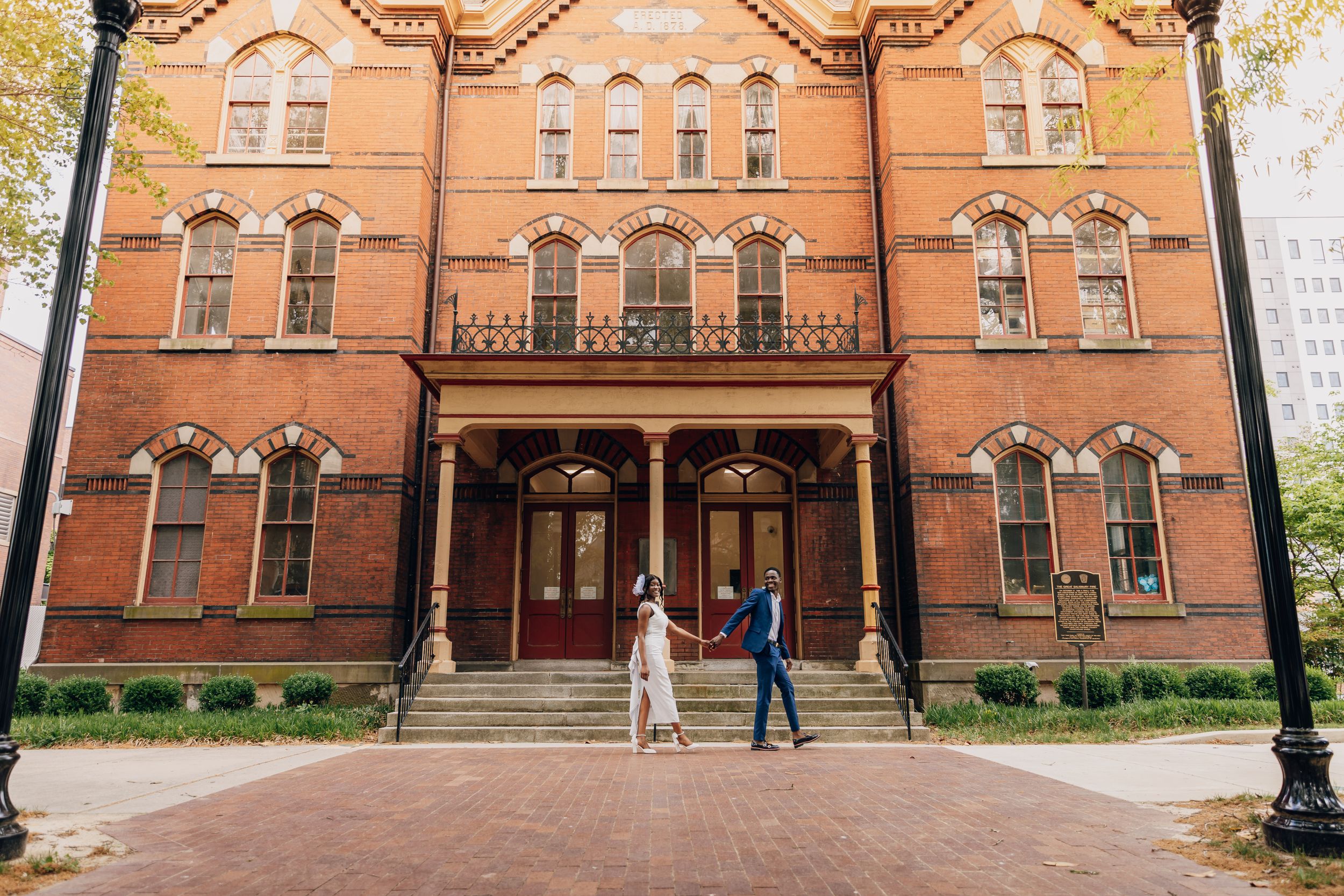 Couple holding hands in front of a historic red brick building with arched windows during an outdoor engagement session.