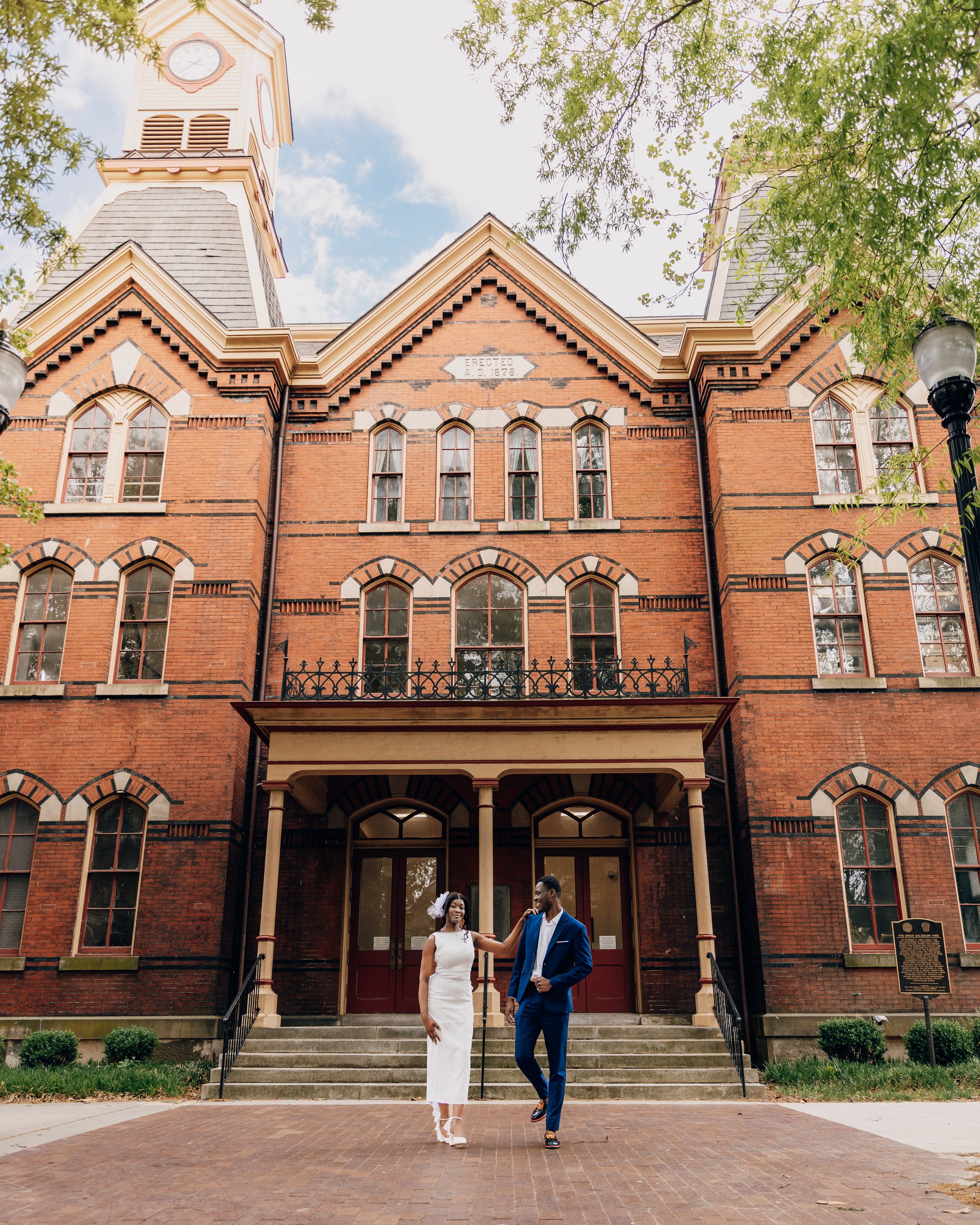 Couple holding hands in front of a historic red brick building with Gothic arched windows on a sunny day.